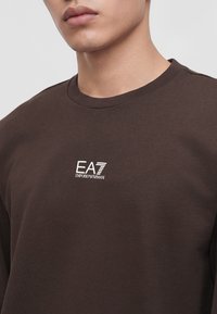Млад човек, носещ тъмнокафява тениска с кръгло деколте и бяло лого EA7 Emporio Armani на гърдите, на светлосив фон.