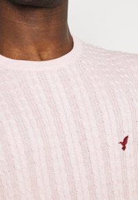 Light pink knitted sweater featuring a textured cable pattern and a small red embroidered logo on the chest. Round neckline.