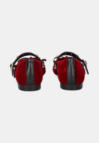 Zapatos de ante rojo con un panel trasero de cuero negro, adornados con tachuelas doradas y una correa ajustable. Suela plana con un patrón de tracción mínimo.
