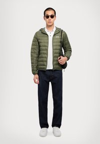 Polstret olivengrønn jakke med hette, hvit polo-skjorte, mørke denimjeans og hvite joggesko. Tilbehøret inkluderer runde, mørke solbriller og en liten veske.