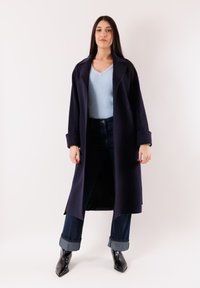 Donna in piedi con un cappotto blu scuro sopra un maglione azzurro, jeans scuri risvoltati e stivaletti neri a punta, su uno sfondo neutro.