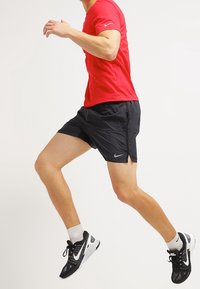 Czerwona koszulka sportowa z krótkim rękawem, w połączeniu z czarnymi szortami. Uzupełniona czarnymi butami do biegania i białymi skarpetkami. W pozycji w pół kroku.