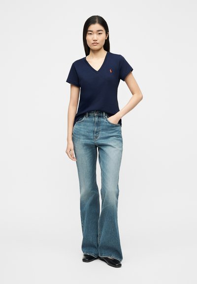 Γυναίκα που φορεί μπλε σκούρο T-shirt με V λαιμόκοψη και μικρό λογότυπο, και ψηλοτάτες φθαρμένες μπλε καμπάνες τζιν με μαύρα παπούτσια, στέκεται με το ένα χέρι στην τσέπη.