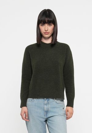 Donkergroen, gebreide trui met een ronde hals en lange mouwen, met een zachte textuur en een iets cropped pasvorm. Gecombineerd met lichtblauwe jeans.