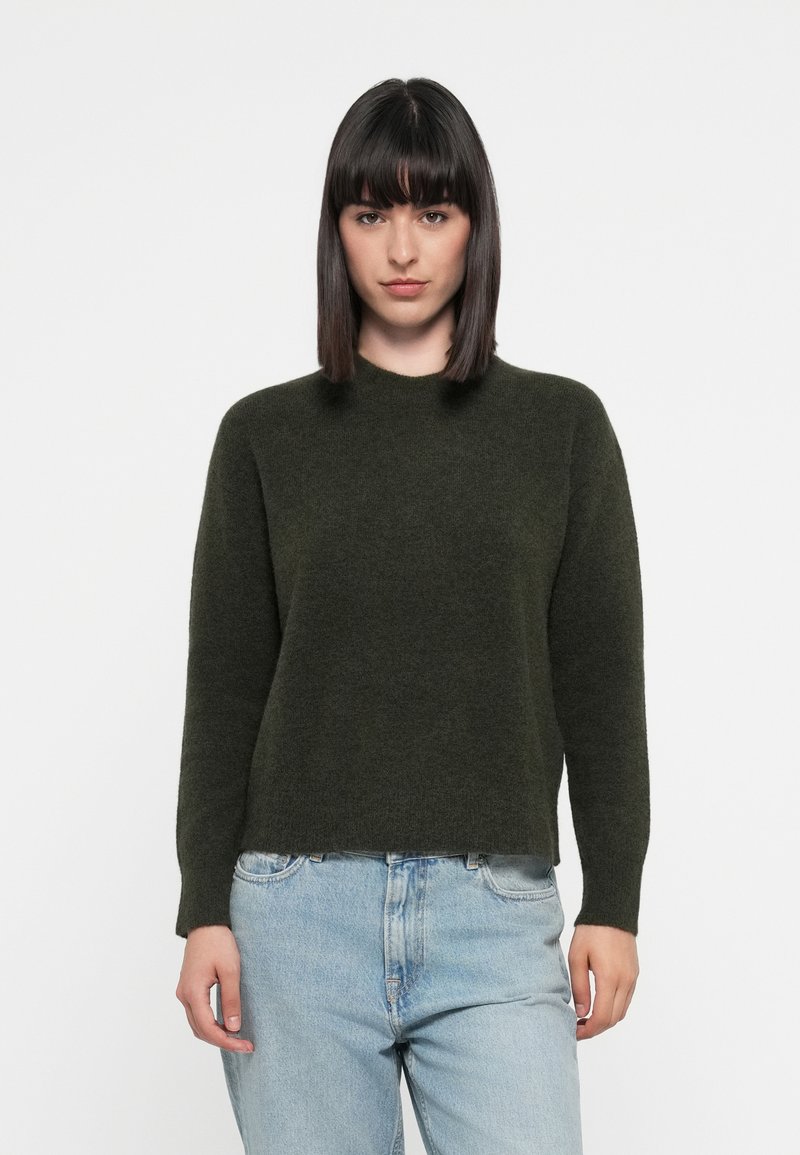 Mørkegrønn, strikket genser med rund hals og lange ermer, som har en myk tekstur og en lett cropped passform. Kombinert med lyseblå jeans.