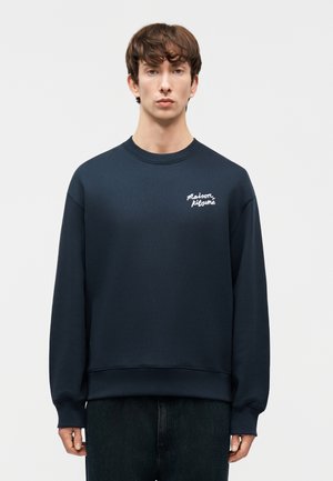 Jeune homme portant un sweat-shirt bleu marine avec le texte blanc "Maison Kitsuné" sur la poitrine, debout devant un fond gris uni.