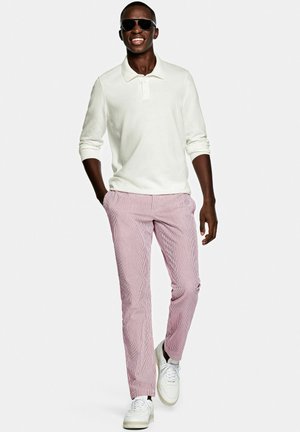 Witte polo met lange mouwen, rood-wit gestreepte broek, witte sneakers en zwarte zonnebril. Staande houding, glimlachend, armen ontspannen.