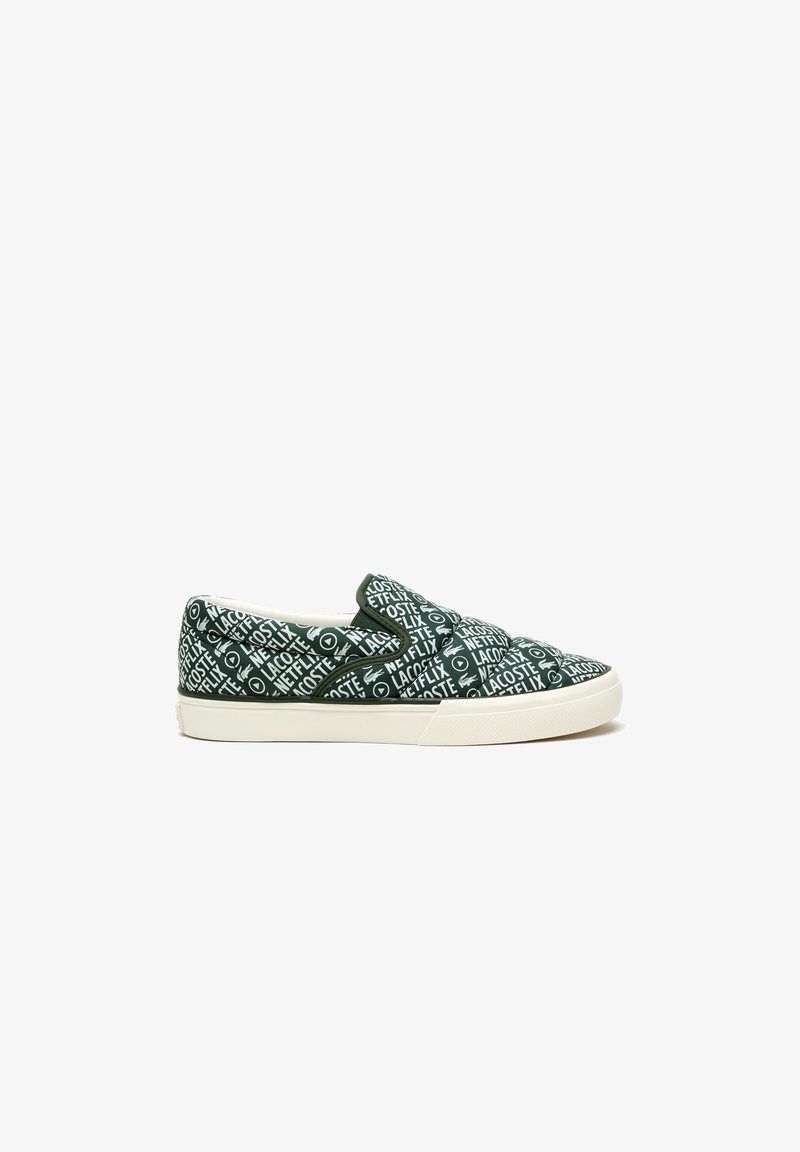 Green slip-on sneakers with white text and patterns, featuring a rubber sole and textile upper. Minimalist design with no visible laces.