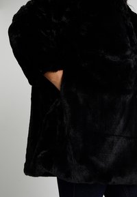 Black faux fur coat with a plush texture, featuring wide sleeves, a high collar, and side pockets for added functionality.