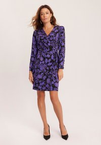 Fioletowa sukienka w kwiaty z długimi rękawami, na czarnym tle, dopasowany krój, gładka tekstura, zestawiona z czarnymi szpilkami i kolczykami w kształcie kół.