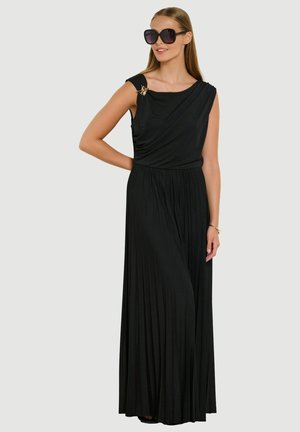 Donna che indossa un lungo vestito nero plissettato senza maniche con una spilla sulla spalla, grandi occhiali da sole neri e un braccialetto d'oro, in piedi su sfondo grigio.