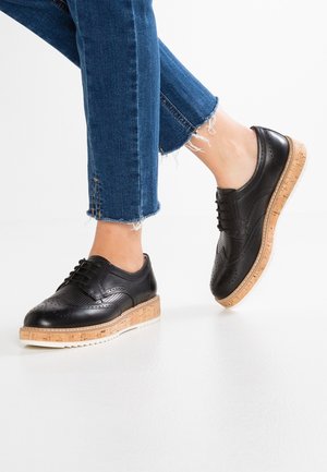 Sorte brogue-sko i læder med korksåler og plateau, båret sammen med blå jeans med frynset kant på en hvid gulv.