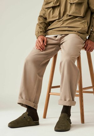 Pullover khaki con polsini elasticizzati, pantaloni beige a risvolto, calze marroni scure, zoccoli in suede verdi, seduto su uno sgabello di legno.