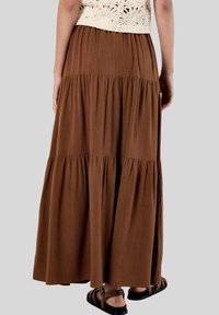 Jupe maxi en couches marron fabriquée en tissu doux avec un design fluide. Dotée d'une taille élastique et accompagnée de sandales à lanières.