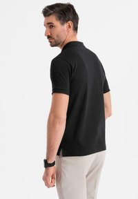 Černé polo tričko s krátkým rukávem, texturovaná tkanina, boční ventily a minimální detaily límce. Nosené s béžovými kalhotami, pohled zezadu.