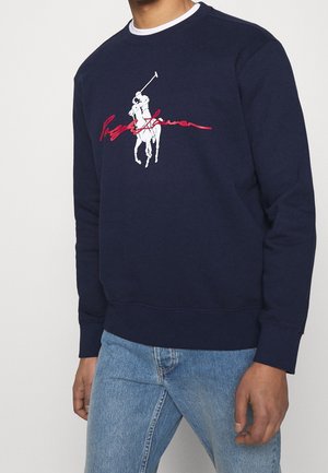 Marineblå sweatshirt med hvid polo-spiller grafik og rød kursiv tekst på brystet, båret med lyseblå jeans.