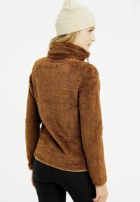 Bruine fleecejack met een hoge kraag en een gestructureerd oppervlak. Gestyld met een getailleerde silhouet, gecombineerd met zwarte leggings en een crèmekleurige beanie.