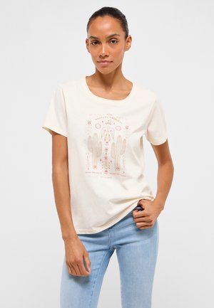 Woman wearing a cream t-shirt with a southwestern cactus and bird graphic, paired with light blue jeans, standing against a white background.