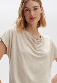T-shirt beige drappe in tessuto morbido e leggero. Presenta una scollatura ampia e maniche corte, con delicate pieghe sulla spalla.