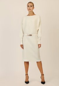 Weißes geripptes Midi-Kleid mit weitem Bootsausschnitt, langen Ärmeln und einem Gürtel mit einer metallischen Schnalle. Schwarze spitz zulaufende Pumps.