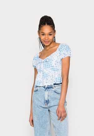 Blusa floral azul clara com mangas bufantes, decote franzido e detalhe de laço, combinada com calças de ganga desbotadas de cintura subida.