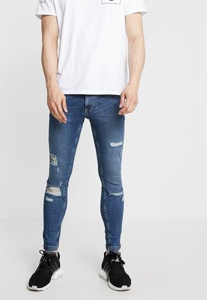 Homme portant un jean slim bleu déchiré, un t-shirt blanc et des baskets noires, debout devant un fond clair uni.