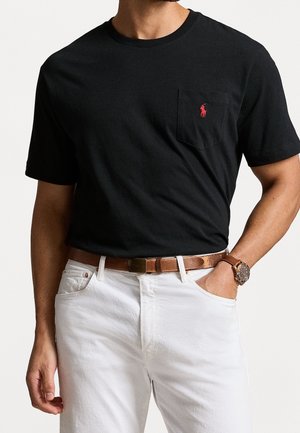 Schwarzes T-Shirt mit kurzen Ärmeln, einer Brusttasche vorne und rotem Logoakzent. Kombiniert mit weißen Jeans und einem braunen Ledergürtel.