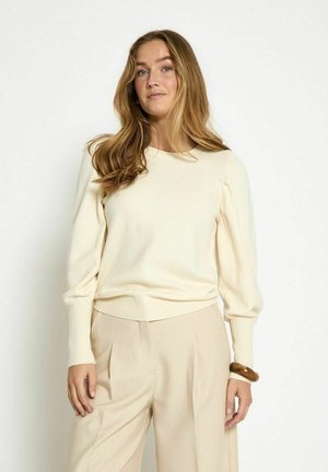 Maglione crema con polsini a coste, una cintura larga e maniche a sbuffo, abbinato a pantaloni beige chiaro.