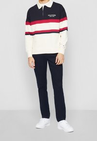 Långärmad polo i marinblått, krämfärgat och rött med horisontella ränder, försedda med en krage och broderad logotyp. Bärs med marinblå byxor och vita sneakers.