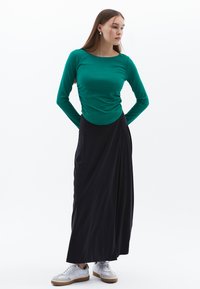 Top verde a maniche lunghe con scollatura rotonda e dettagli arricciati, abbinato a una gonna maxi nera fluida e sneakers bianche con suole marroni.