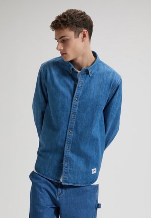 Młody mężczyzna z kręconymi włosami, ubrany w niebieską dżinsową koszulę i dżinsy, patrzący w dół w lewo na jednolitym tle.