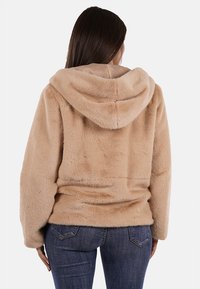 Giacca in pelliccia sintetica color beige chiaro con cappuccio, caratterizzata da una texture morbida e una vestibilità rilassata, vista da dietro. Abbinata a jeans denim scuri.