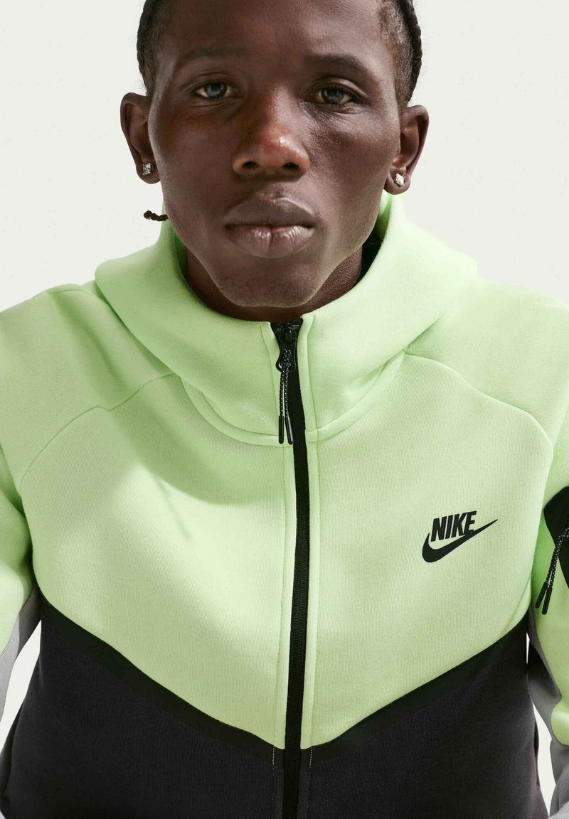 Nærbillede af en person iført en lysegrøn og sort Nike lynlåstrøje med høj krave, der kigger direkte frem med et neutralt udtryk.