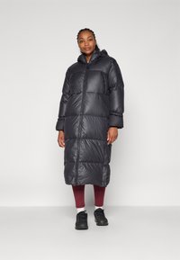 Svart pufferjacka med lång design, utrustad med huva, dragkedja fram och breda horisontella kviltade sektioner. Texturerad tyg med en glansig yta.