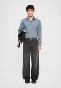 Chemise à rayures bleu clair, boutonnée ; jean gris foncé ample ; chaussures noires ; tenant un sac noir. Design simple, style décontracté.