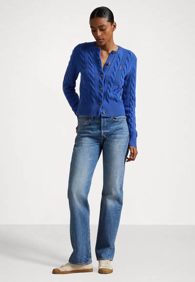 Blauwe kabelgebreide cardigan met een ronde hals, houten knopen en geribbelde manchetten, gecombineerd met lichtblauwe recht gesneden jeans en beige sneakers.