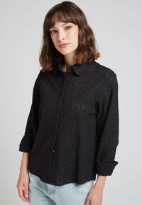 Chemise noire à boutons avec un motif texturé, manches longues retroussées au niveau des poignets, et col. Comprend des détails découpés subtils.
