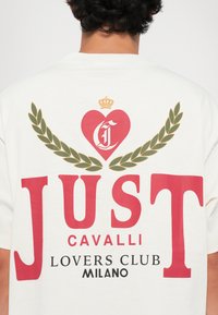 White cotton t-shirt featuring a large red text "JUST," surrounded by green laurel leaves and a red heart with a crown above it.