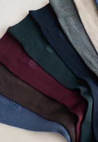 Sette paia di calzini di lana piegati in beige, grigio, blu navy, verde scuro, bordeaux, marrone e blu, disposti diagonalmente su una superficie chiara.