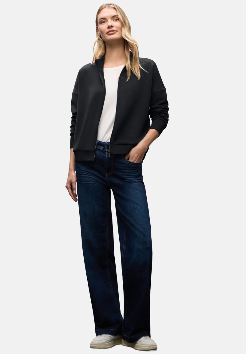 Chaqueta negra con cremallera sobre una blusa blanca, combinada con jeans de pierna ancha de color azul oscuro y zapatillas blancas. Tejido suave, corte relajado y diseño informal.