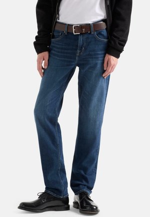 Man draagt donkerblauwe spijkerbroek, zwarte leren nette schoenen, bruine riem, wit overhemd en zwarte jas, staand tegen een witte achtergrond.