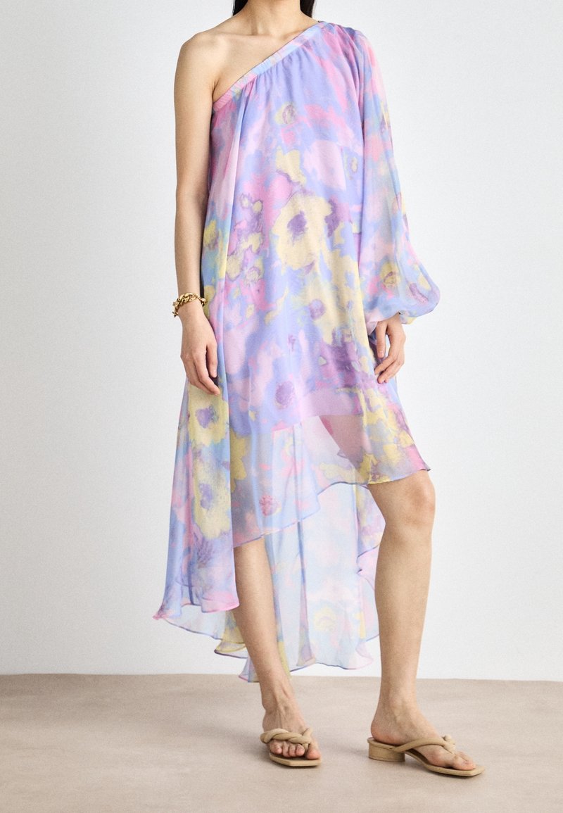 Femme portant une robe pastel fluide asymétrique à une épaule avec des motifs floraux, associée à des sandales beige à bout ouvert.