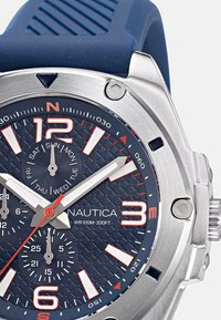 Reloj Nautica con correa de goma azul marino, bisel de acero inoxidable, esfera texturizada en azul oscuro, marcadores de hora en blanco y naranja, y subesferas.