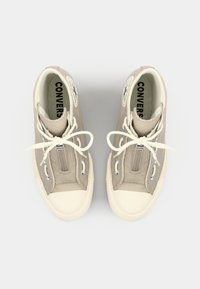 Beige lerrets høye sneakers med hvite gummisåler, sølv glidelåser og hvite snøringer. Har en sirkulær logo-patch på hver ytterside.