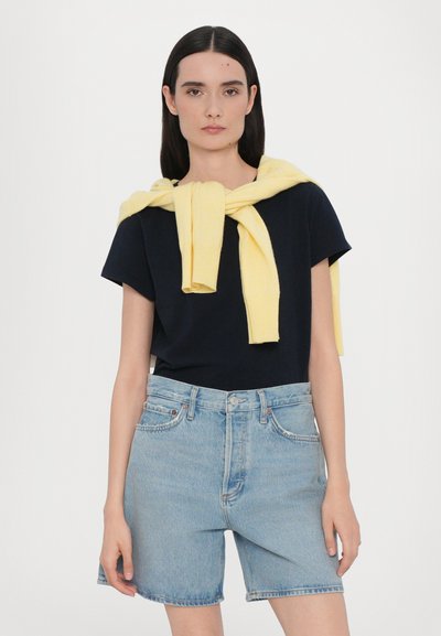 Μπλε ναυτικό μπλουζάκι συνδυασμένο με ανοιχτό μπλε τζιν σορτς. Ένα κίτρινο πουλόβερ είναι περασμένο πάνω από τους ώμους, με ευθείες γραμμές και ριμπ ύφανση.