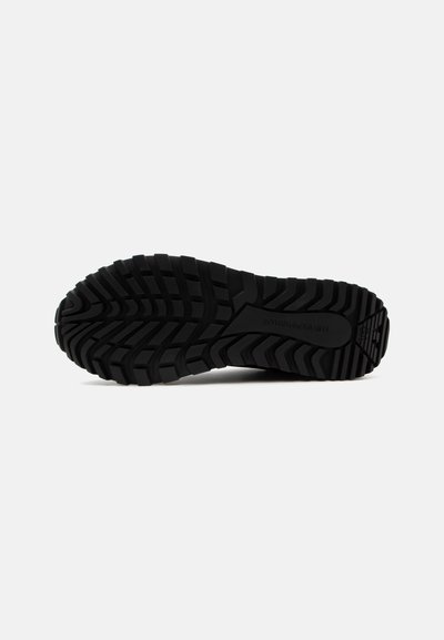 Semelle de chaussure en caoutchouc noir avec un motif de bande de roulement profond et rugueux pour la traction, présentée sur un fond clair uni.