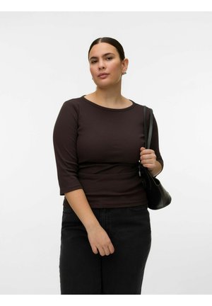 Femme aux cheveux foncés tirés en arrière, portant un haut noir à manches trois-quarts et un pantalon noir, tenant un sac à bandoulière noir.