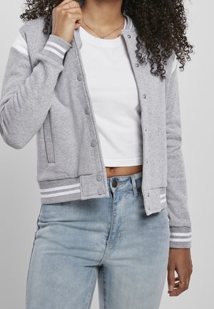 Personne portant une veste varsity grise à boutons-pression par-dessus un crop top blanc et un jean taille haute bleu clair, avec des cheveux bouclés visibles.