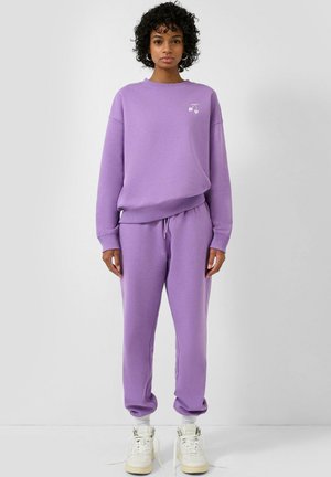 Femme aux cheveux bouclés portant un sweat-shirt et un pantalon de survêtement lavande assortis, des chaussettes blanches et des baskets montantes blanches, debout devant un fond uni.