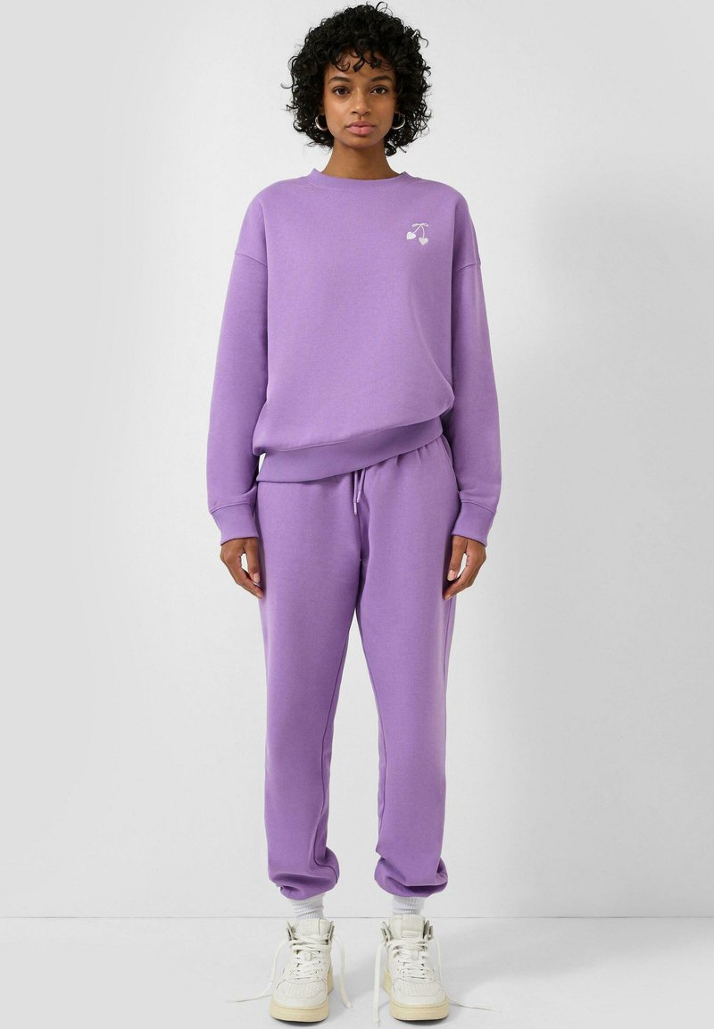 Frau mit lockigem Haar, die einen passenden lavendelfarbenen Sweatshirt und Jogginghose trägt, weiße Socken und weiße High-Top-Sneakers, stehend vor einem schlichten Hintergrund.